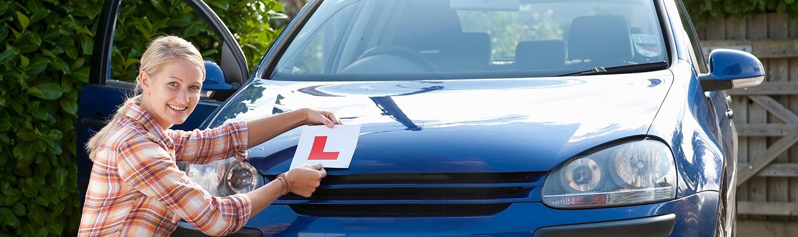 Happy young blonde woman kneeling by car applying L plates. Pleased she has used Zixty for her learner driver insurance.